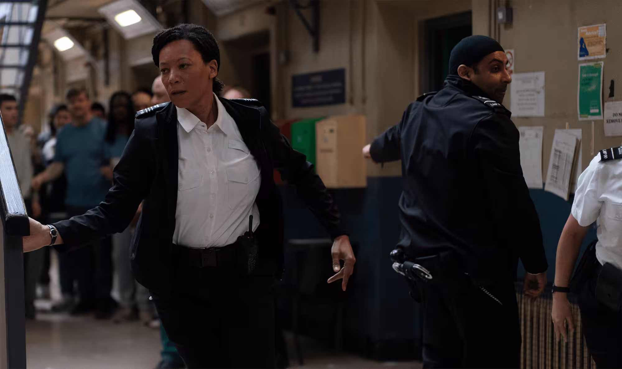 Female officer running through a busy cell block as other guards restrain prisoners.