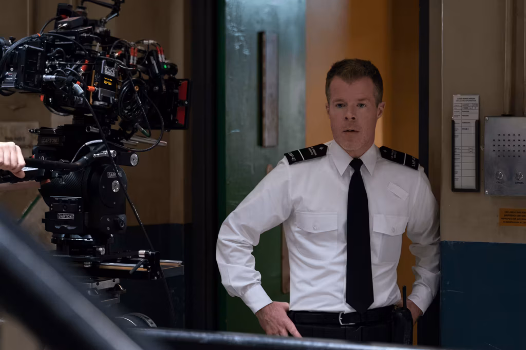 Man acting as a prison officer looking concerned, with a camera visible at the edge of the frame.