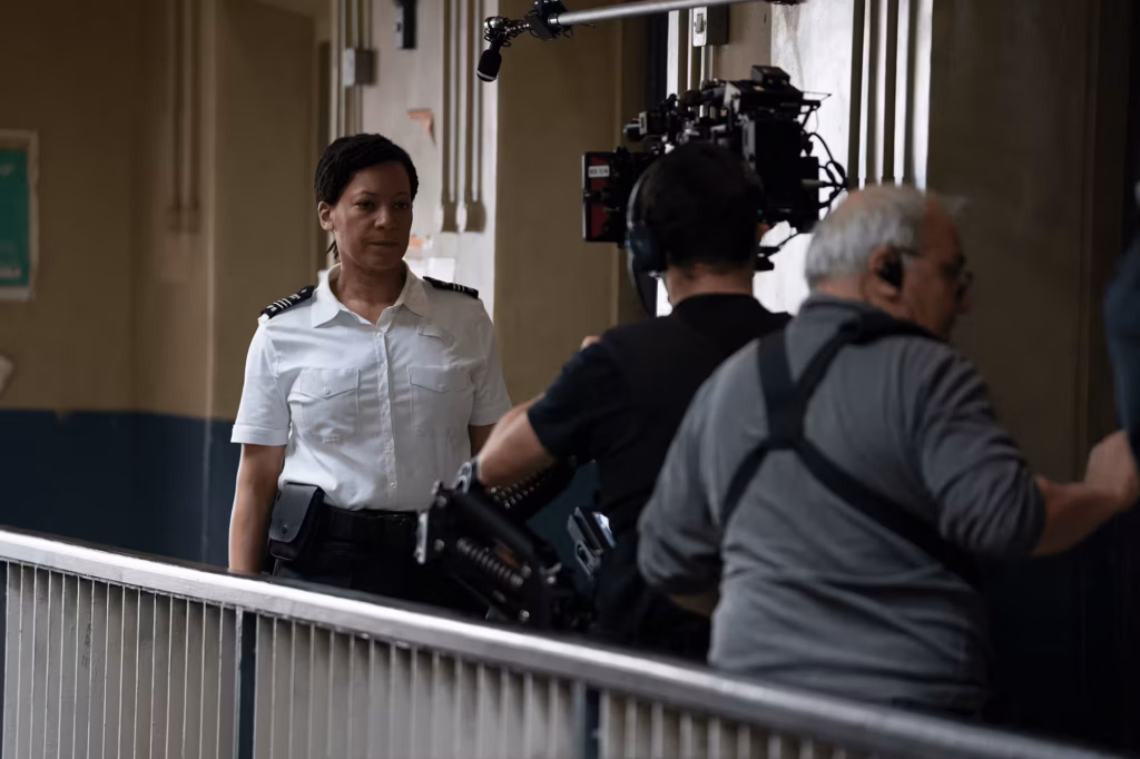 Woman in a prison officer outfit, with production crew and camera equipment preparing to shoot in the background.