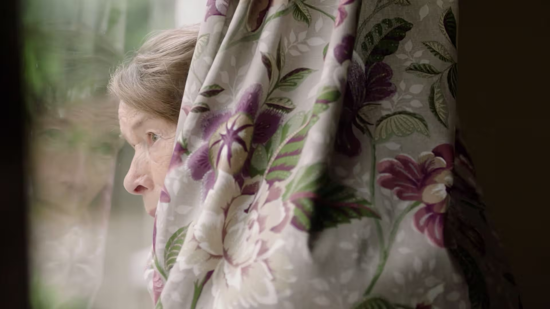 Elderly woman hiding behind a curtain while looking out through a window.