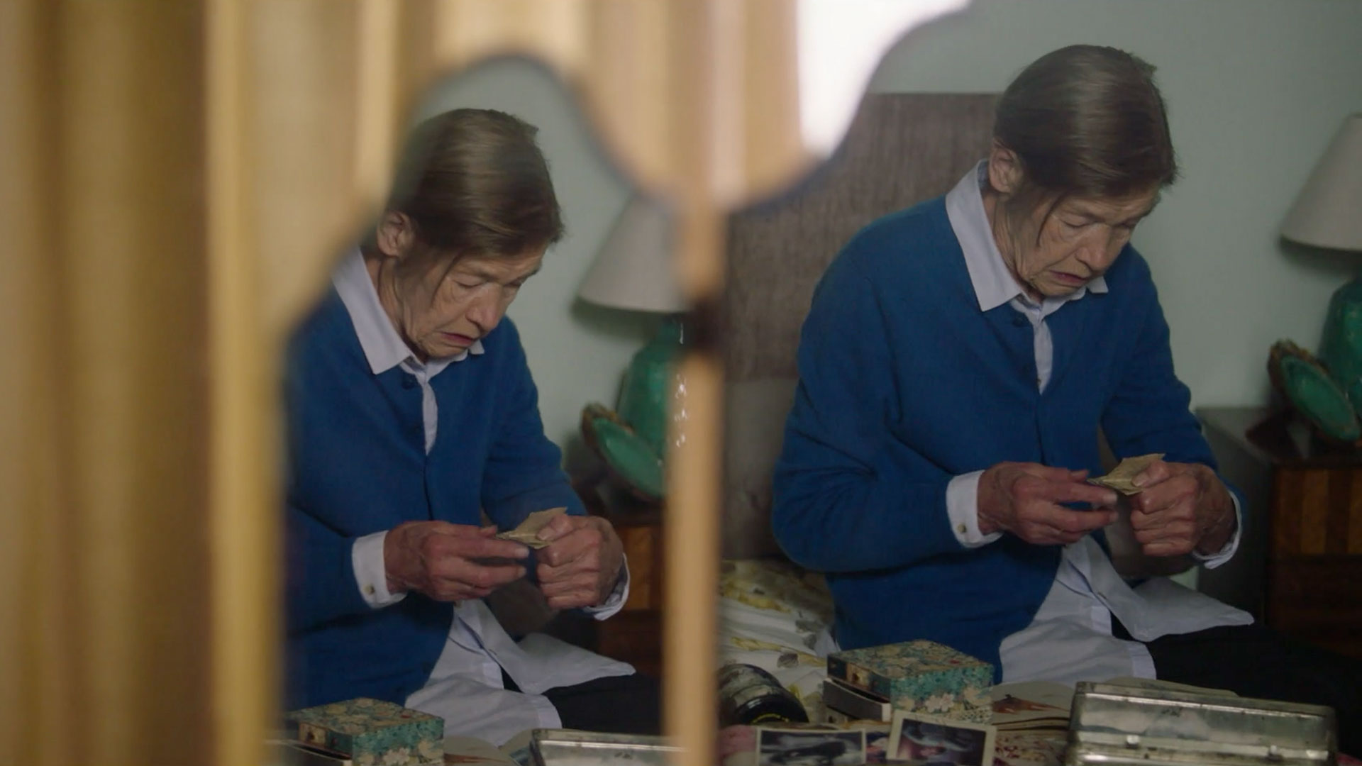 Reflection of an elderly woman in a mirror as she looks through newspaper cuttings and photographs.