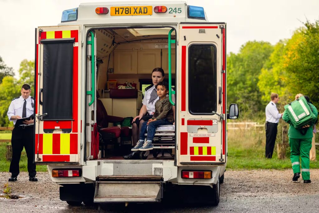 Ambulance scene with a female police officer and a child seated inside, while paramedics and police officers work outside the vehicle.