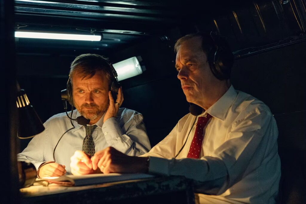 Two man wearing headsets sit in a dimly lit control booth, focused on monitoring a witness.