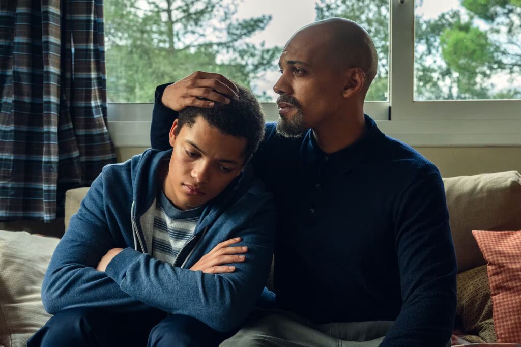 An adult comforts a sad-looking teenager on a couch, resting a hand on their head while they sit together near a window.